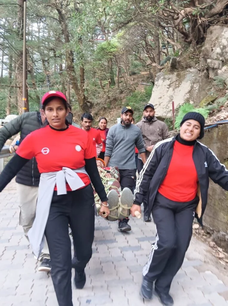 Two AapdaVEER women volunteers leading a stretcher carry up a pine-forested path during an AapdaABHYAS preparedness drill